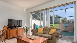 A living room with a TV, wooden furniture, a sofa with cushions, and a view of a swimming pool.