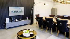 A modern dining area with a long table, dark upholstered chairs, a glass-top coffee table with decorative items, a dark wood panel wall, and a mounted artwork. - Natree Pool Villa Pattaya (Pattaya)