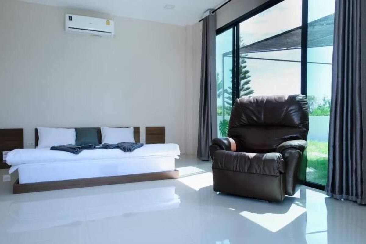 A modern hotel room with a large bed, a leather chair, and a view of greenery through large windows.