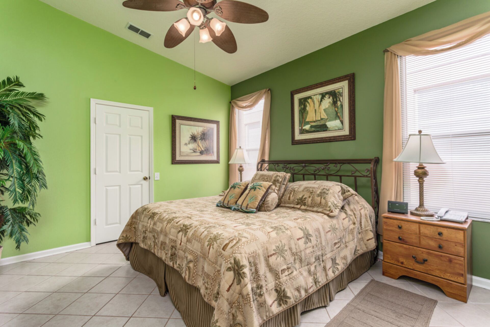 A bedroom with a bed, a nightstand, a lamp, a ceiling fan, a door, and two framed pictures on the wall.