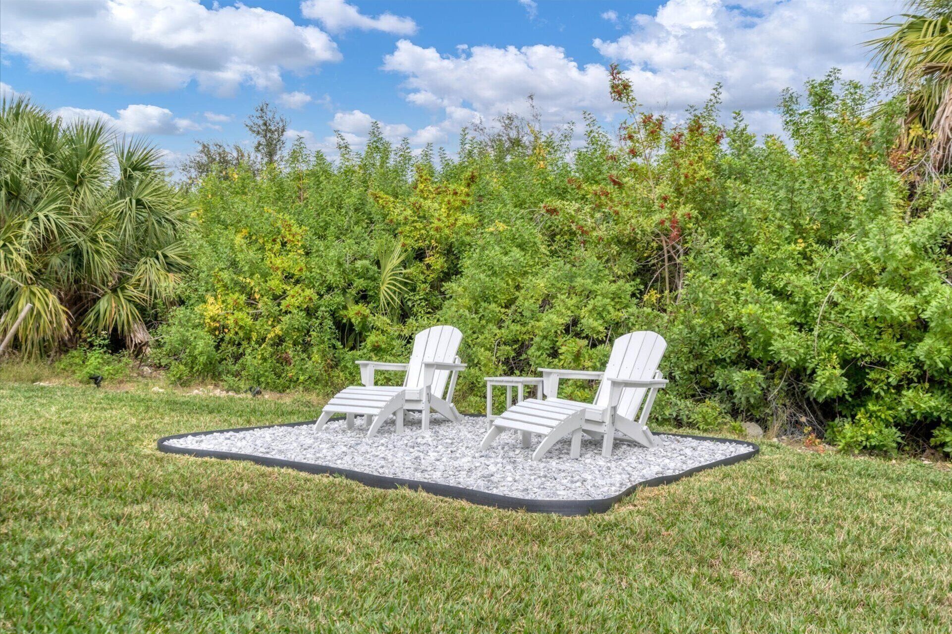 A garden area with two white Adirondack chairs and a small table, surrounded by a gravel border, set against a backdrop of lush greenery and a clear blue sky.