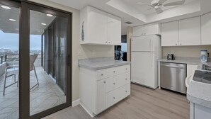 A modern kitchen with white cabinets, a stainless steel refrigerator, and a dishwasher. There's a balcony with a glass door and a ceiling fan. - Stay at Sarasota County Villa 1005: Gulf Views & Resort Amenities (Sarasota County)