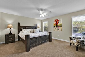 Ein Schlafzimmer mit Bett, Nachttischen, Deckenventilator, Fenster mit Jalousien, einem Sessel und einem Bild an der Wand.