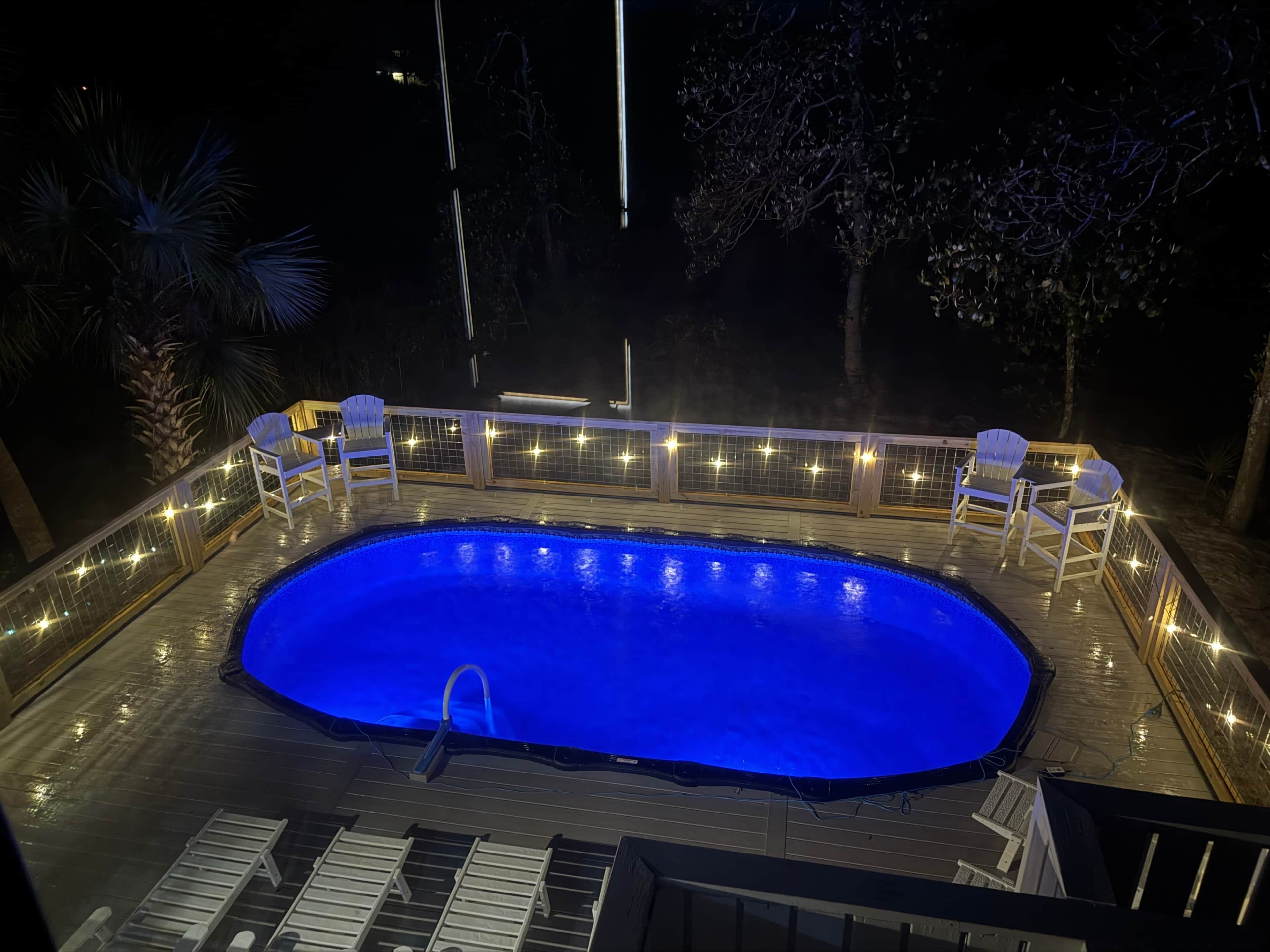 A swimming pool with a wooden deck, surrounded by chairs and string lights.