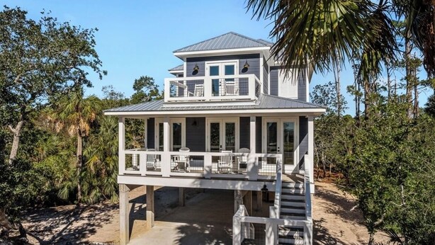 A two-story beachfront house with a balcony, surrounded by palm trees and a sandy beach.