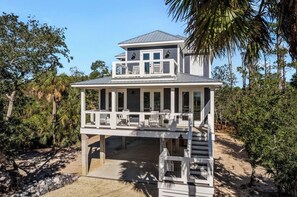 Una casa fronte spiaggia su due piani, con un balcone, circondata da palme e una spiaggia sabbiosa.