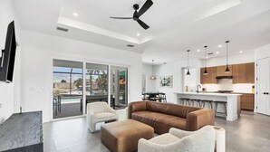A modern living room with a brown leather sofa, a white armchair, and a beige ottoman. There is a dining area with a table and chairs, and a kitchen with wooden cabinets and pendant lights. - Charlotte County Villa 5892 - Pool Home Retreat (Charlotte County)