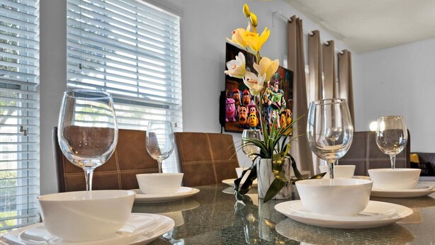 Une table à manger dressée avec de l’assiette blanche, des bols et des verres à vin, agrémentée d’un vase de fleurs jaunes et d’un tableau coloré en arrière-plan.