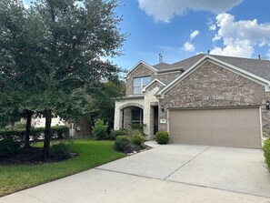 A two-story house with a stone facade, a garage, and a well-maintained front yard with trees and shrubs. - Charming house in The Woodlands - Creekside (Tomball)