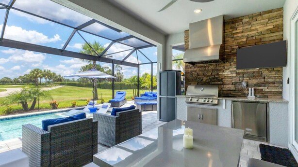 A modern outdoor kitchen with a stone backsplash, a large window overlooking a pool and greenery, and comfortable seating by the pool. - Premier Golf & Pool Retreat – Charlotte County Villa 5902 (Charlotte County)