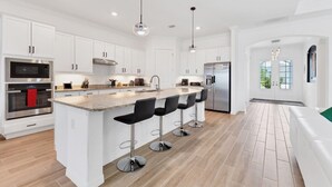A modern kitchen with white cabinetry, stainless steel appliances, a central island with bar stools, and a view to the outside. - Premier Golf & Pool Retreat – Charlotte County Villa 5902 (Charlotte County)