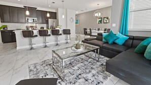 Ein modernes Wohnzimmer mit einem Glastisch, einem dunkelgrauen Sofa mit türkisfarbenen Kissen und einer Küchenzeile mit dunklen Schränken und Edelstahlgeräten.
