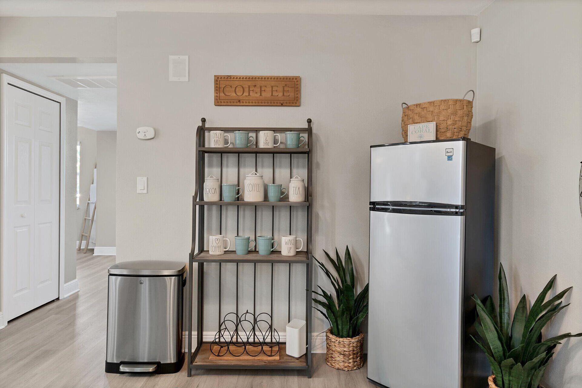 A kitchen with a stainless steel refrigerator, a coffee shelf, a trash bin, and two potted plants.