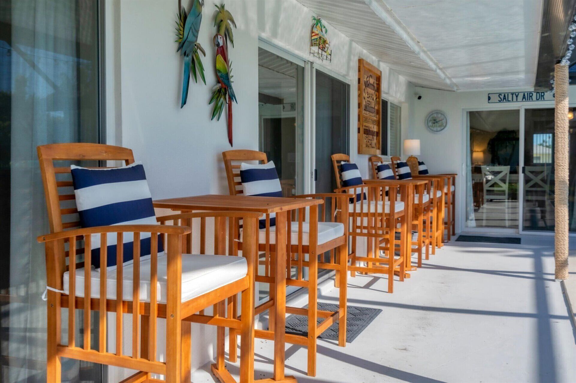 A row of wooden chairs with white cushions and blue and white striped pillows, arranged on a veranda with a white ceiling and a sign reading "SALTY AIR DR" in the background.
