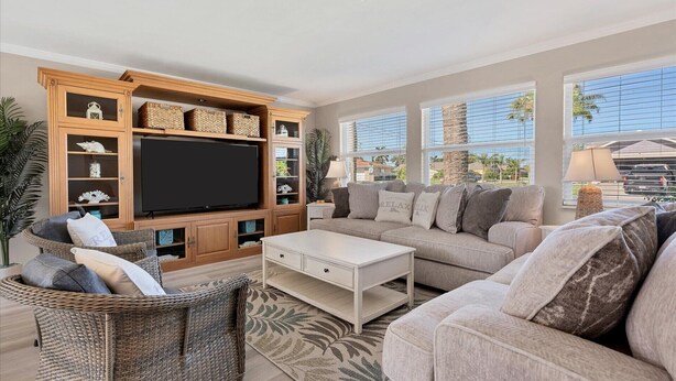 A living room with a large flat-screen TV, wicker furniture, a white coffee table, and a view of palm trees and a building through large windows.