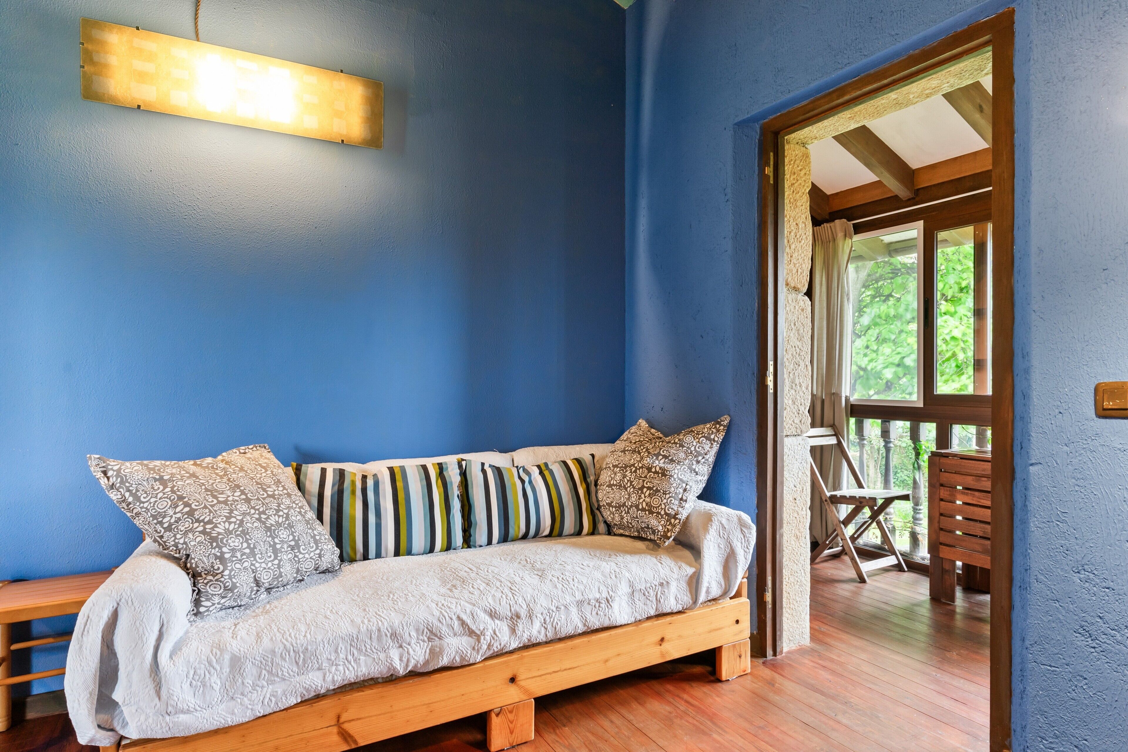 Un sofá de madera con cojines estampados, un banco y una lámpara colgante en una habitación con paredes azules y suelo de madera.