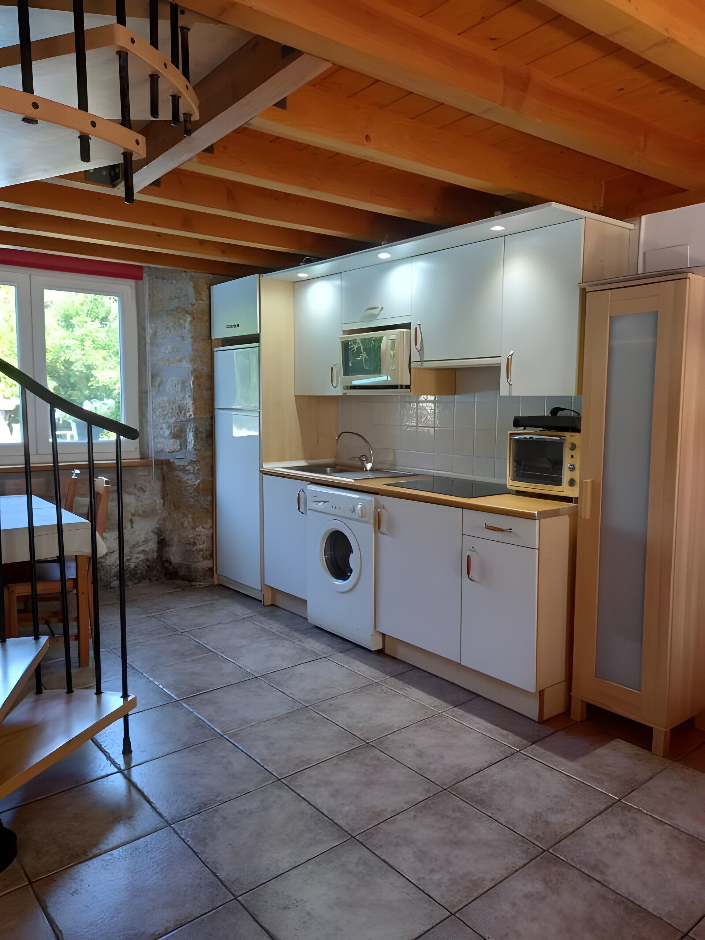 Een praktische keuken met een wasmachine, magnetron en oven, houten balken in het plafond, en een eethoek met een tafel en stoelen.