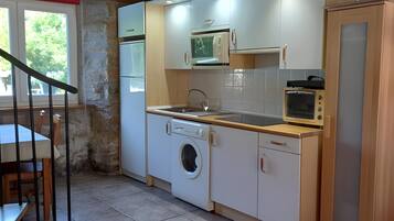 Een praktische keuken met een wasmachine, magnetron en oven, houten balken in het plafond, en een eethoek met een tafel en stoelen.