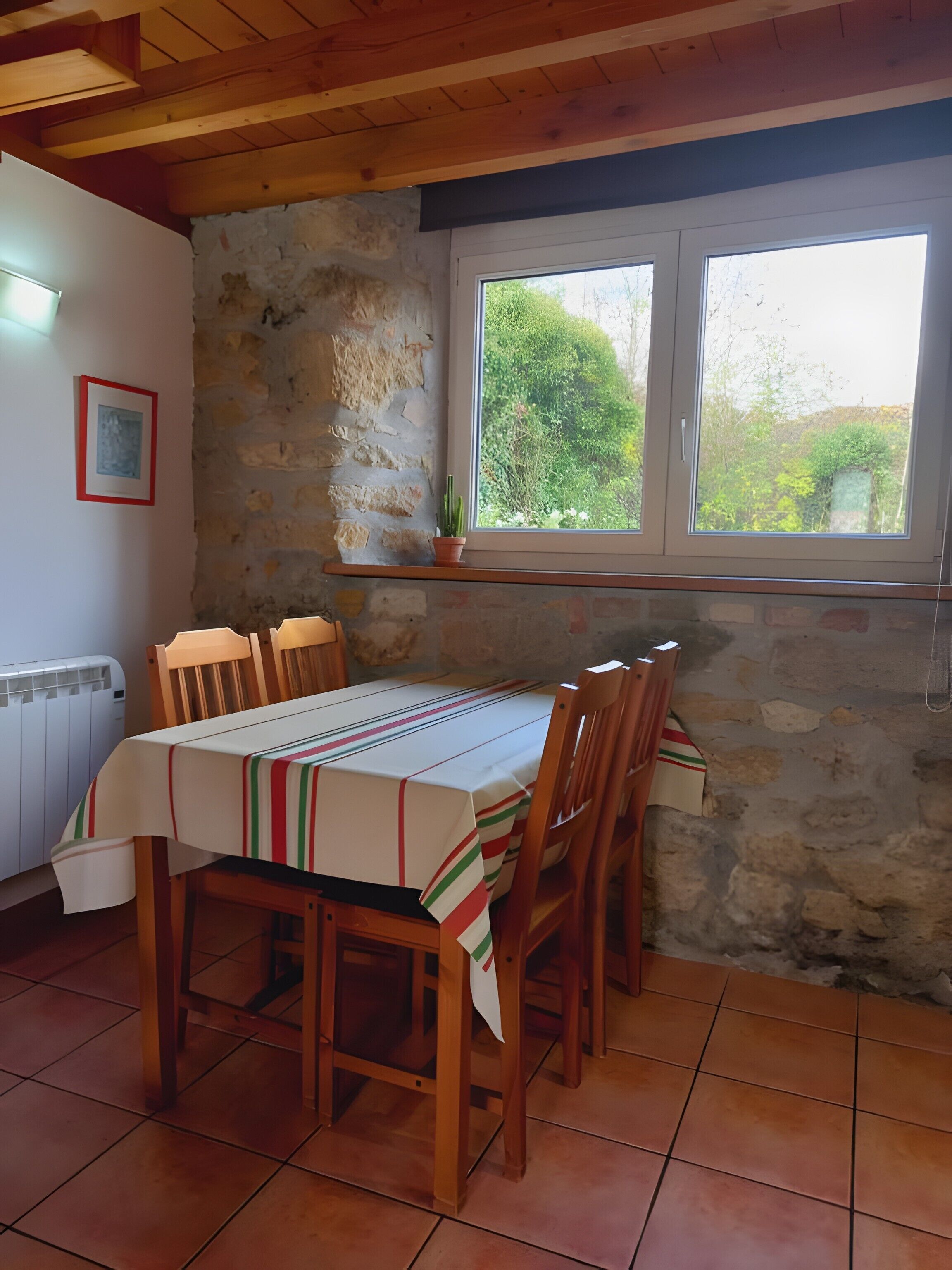 Un coin repas avec une table dressée pour quatre personnes, des chaises en bois, un mur en pierre, une fenêtre donnant sur la verdure et un radiateur.
