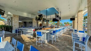 An outdoor dining area with multiple tables and chairs, a bar counter, and a pool in the background.