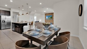 A modern kitchen with a central island, stainless steel appliances, pendant lighting, and a dining area with a long table set for four.