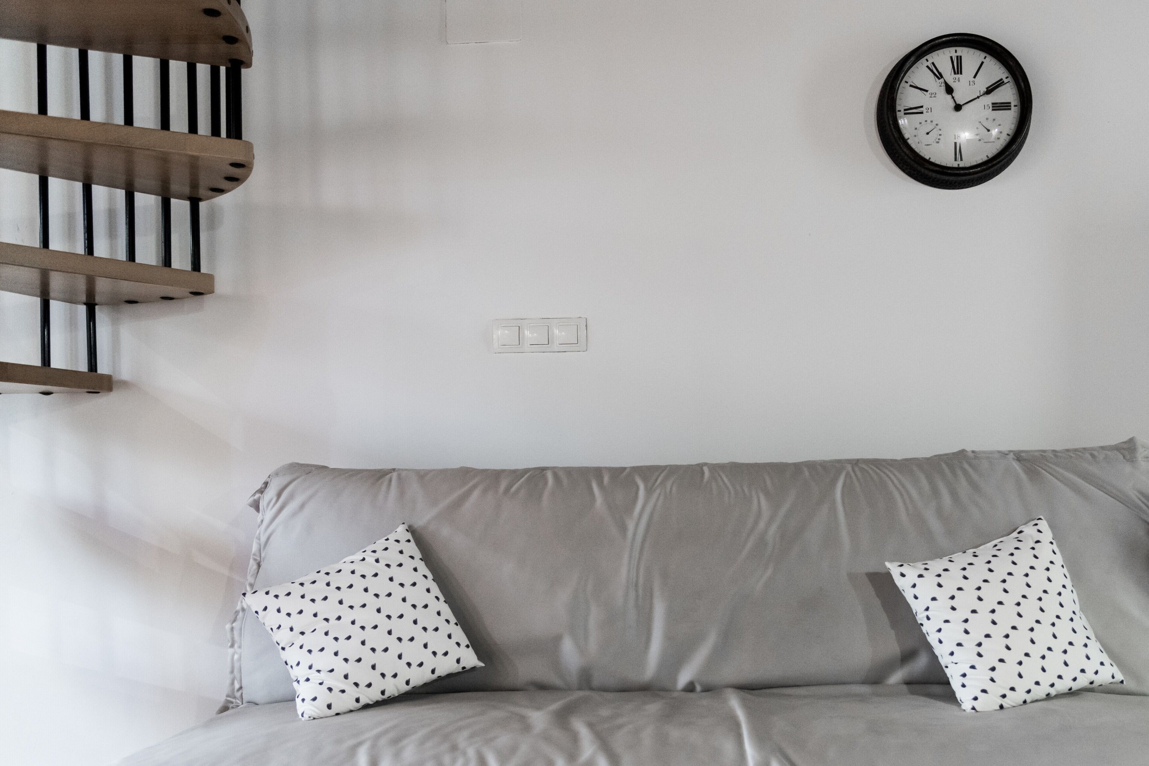 Una habitación con un sofá gris, dos almohadas estampadas, un reloj de pared y una estantería de madera.
