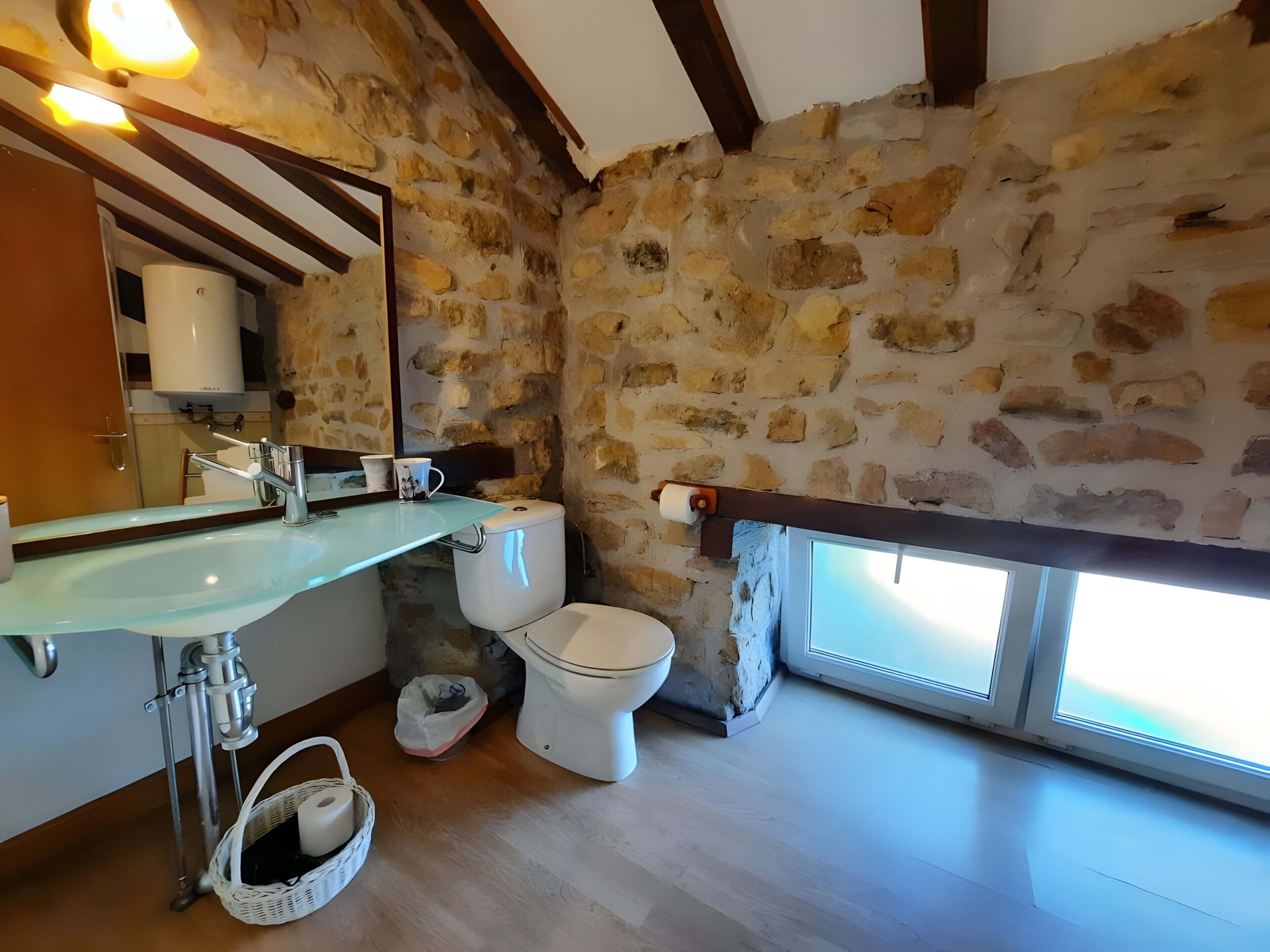 Un baño con pared de piedra, lavamanos de vidrio, inodoro y ventana.