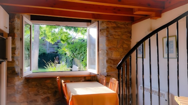 Una habitación con pared de piedra, techo de madera, ventana con vista a follaje, escalera con barandillas negras y una mesa con mantel blanco.