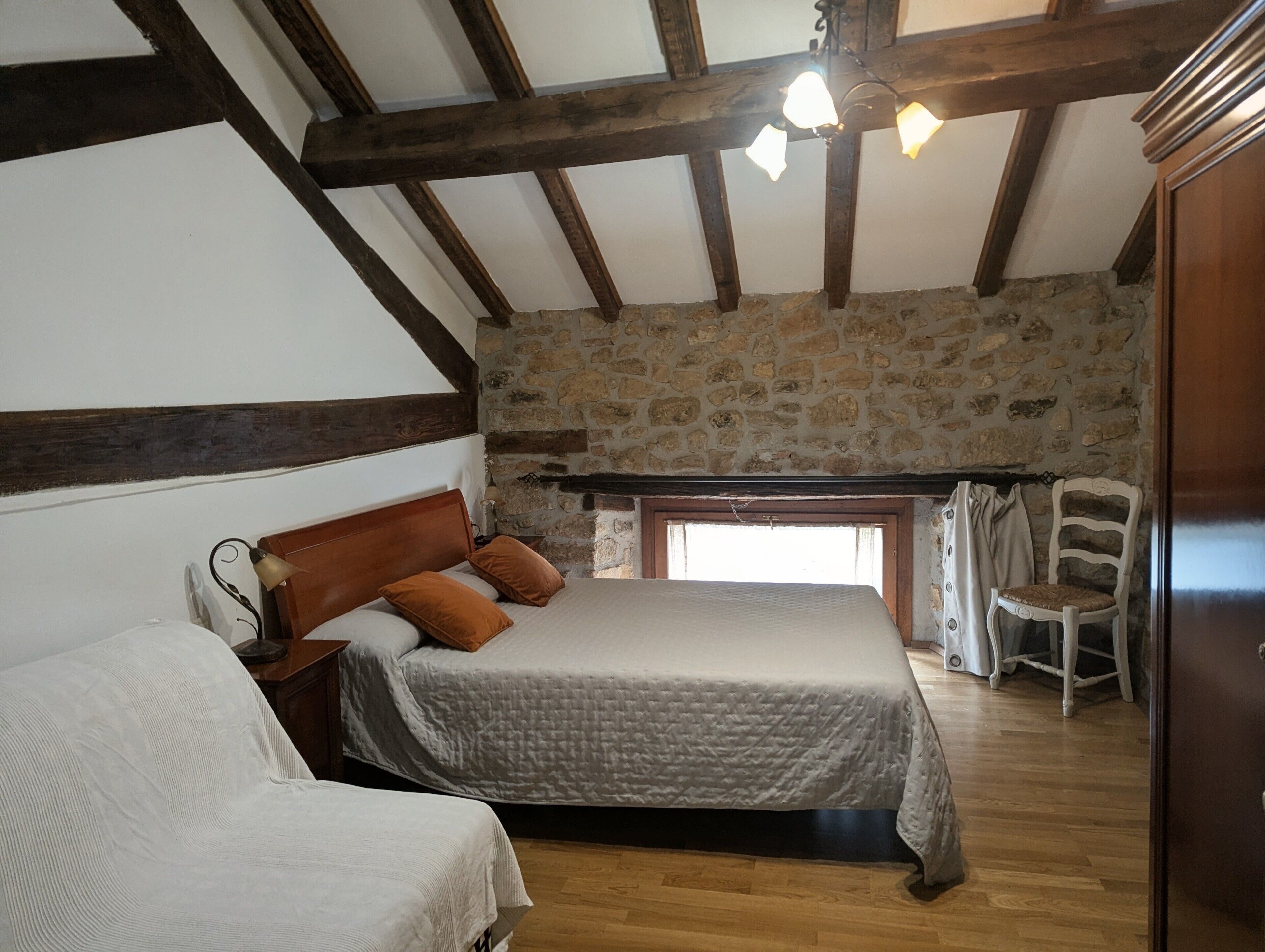 A bedroom with a large bed, a smaller bed, a stone wall, wooden beams, and a window.
