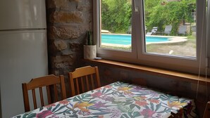 A dining area with a table covered in a floral tablecloth, wooden chairs, a stone wall, a window with a view of a swimming pool, and a white refrigerator. - Apartment in Alsasua with Shared Pool, Wi-Fi and Air Conditioning (Alsasua)