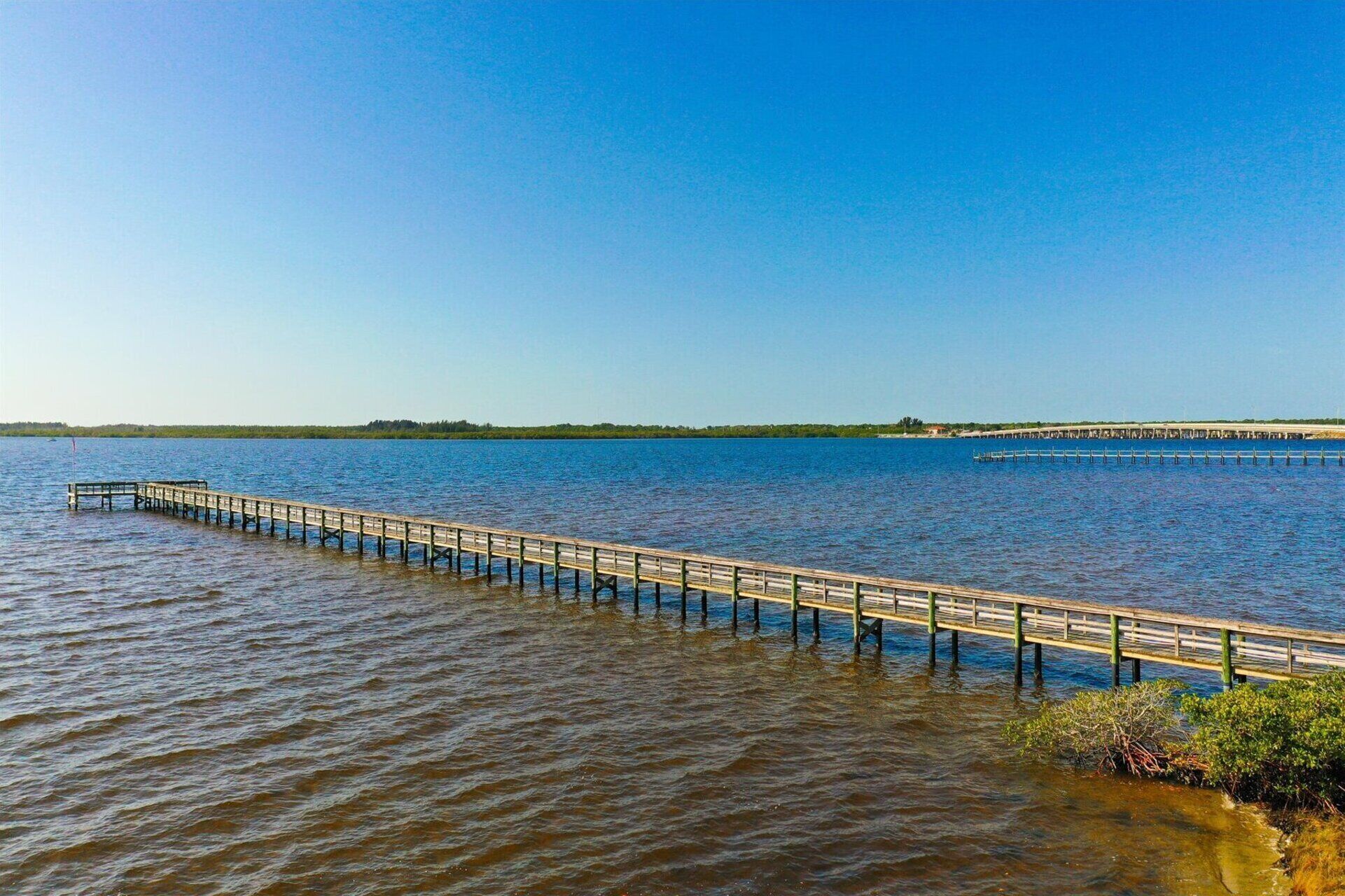 A wooden pier extending into a calm body of water, with a clear sky above and a distant shoreline visible.