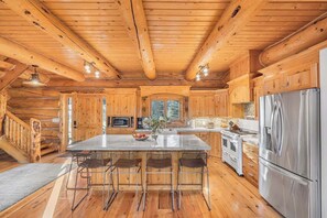 L’intérieur d’une cabane en rondins est aménagé avec un îlot central, des tabourets de bar, un réfrigérateur en acier inoxydable et une cheminée.
