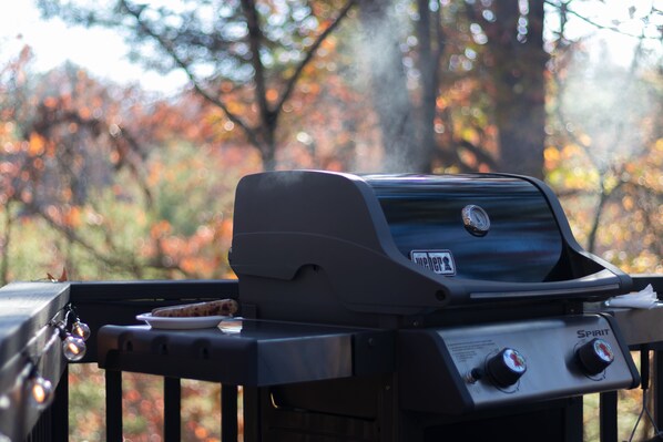 A Weber Spirit grill with a plate of food on it, set against a backdrop of autumn foliage.