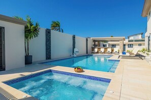 Una piscina con borde de azulejos, una zona de estar con tumbonas y un edificio moderno con balcón.