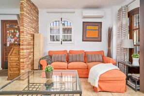 A living room with a brick wall, a glass coffee table, an orange sofa, a white throw blanket, and a bookshelf.