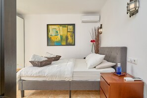 A modern bedroom with a grey bed, white bedding, a wall-mounted air conditioner, and a painting.