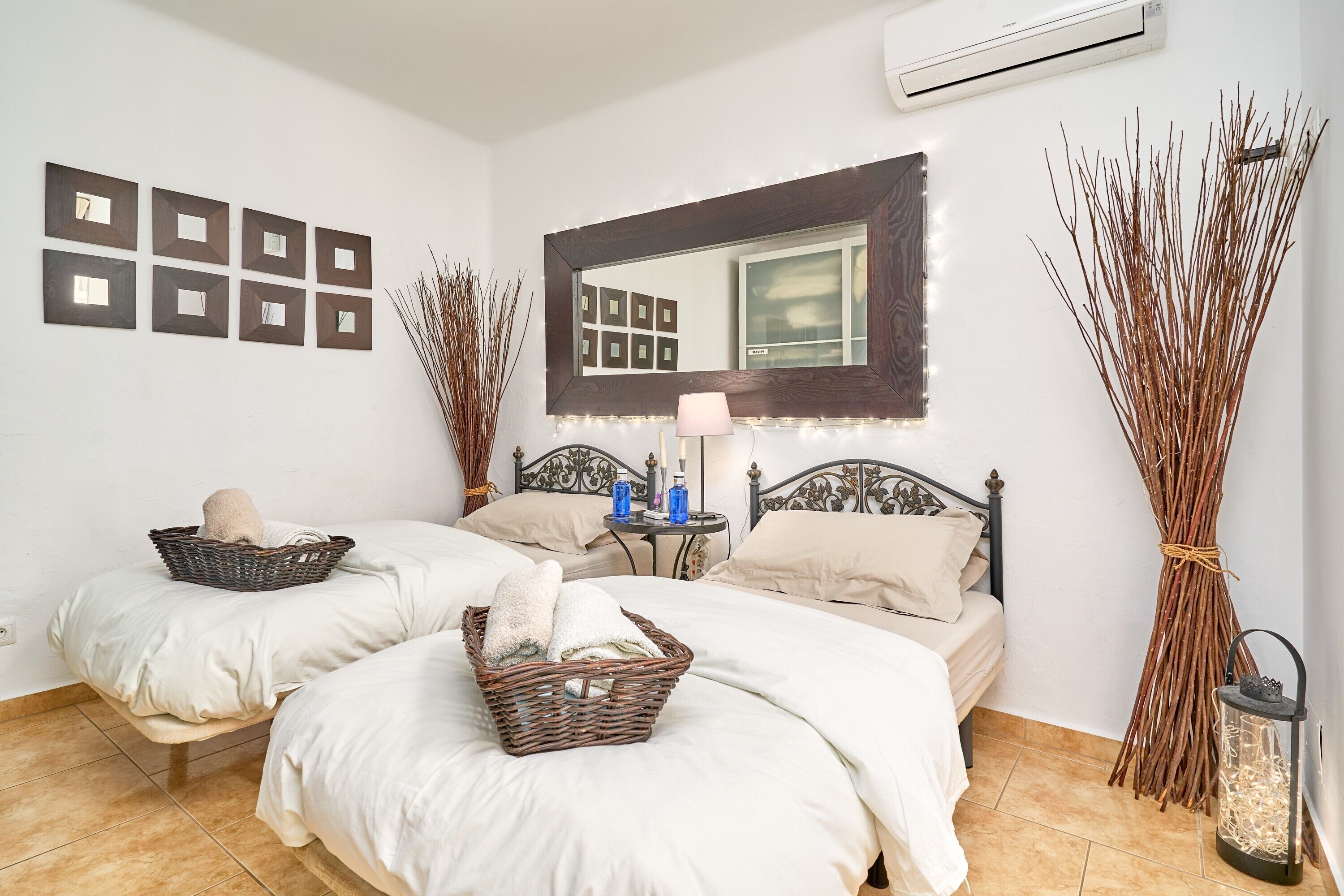 A bedroom with two beds, a mirror, wall-mounted picture frames, a bedside table with a lamp, and decorative branches.