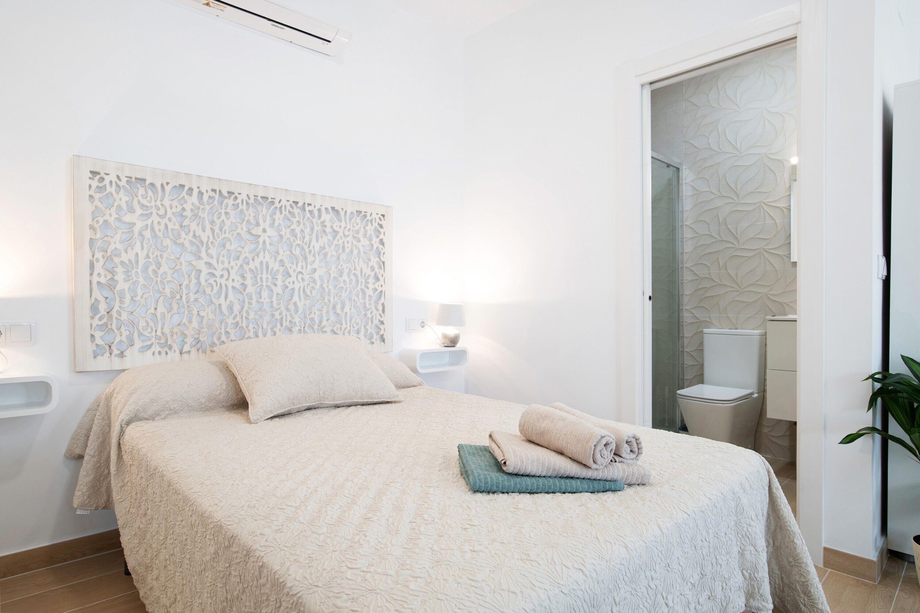 Une chambre avec un grand lit, une lampe fixée au mur, une table de chevet et une salle de bain visible à travers une porte ouverte.