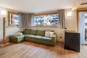 A room with a green sectional sofa, wooden flooring, a black cabinet, and a view of buildings through large windows. - Apartment 'Gavia Bormio' with Mountain View, Balcony and Wi-Fi (Bormio)