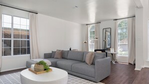 Een moderne woonkamer met een grijze bank, een ronde witte salontafel en een groot raam met gordijnen.