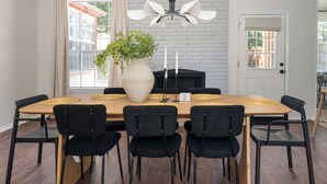 Een eetruimte met een lange houten tafel, zwarte stoelen, een grote vaas met planten, een wit bakstenen haard en een plafondlamp.