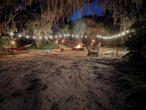 Un espacio al aire libre con arena, una hamaca, sillas, un hogar para fuego y luces colgantes.