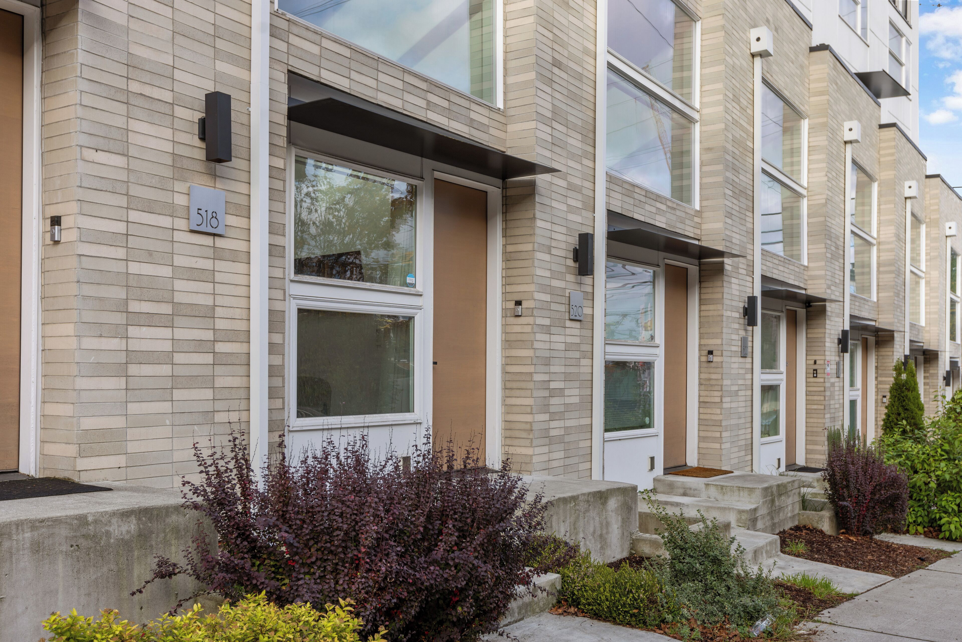 A row of modern townhouses with numbered doors, a brick facade, and a small front yard with shrubs.