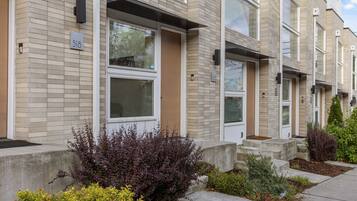 A row of modern townhouses with numbered doors, a brick facade, and a small front yard with shrubs.