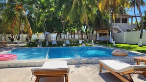 Une piscine avec des chaises longues, des palmiers et un petit chalet à toit de chaume portant l’inscription « Rancho Niebella ».
