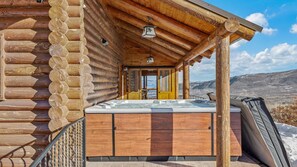 A log cabin with a hot tub, wooden exterior, and a view of a snowy landscape.