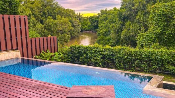 An infinity pool with a wooden deck, surrounded by lush greenery and a river view. - Royale Riviera Resort Private Pool Villas (Langkawi)