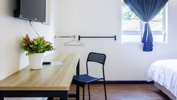 A room with a wooden table, a black chair, a potted plant, a TV mounted on the wall, a window with curtains, and a bed.