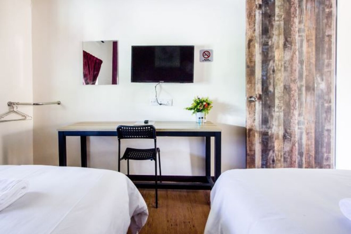 Habitación de hotel con dos camas, un escritorio con una silla, un televisor montado en la pared, una planta pequeña en el escritorio y una puerta de madera.