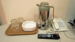 A wooden tray with a white bowl, three glass jars, a silver coffee pot, a white telephone, and a remote control on a white surface.